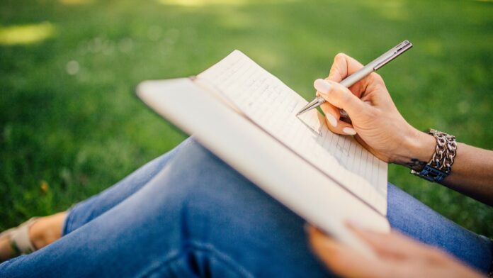 Personne assise dans l'herbe qui écrit avec un stylo dans un carnet